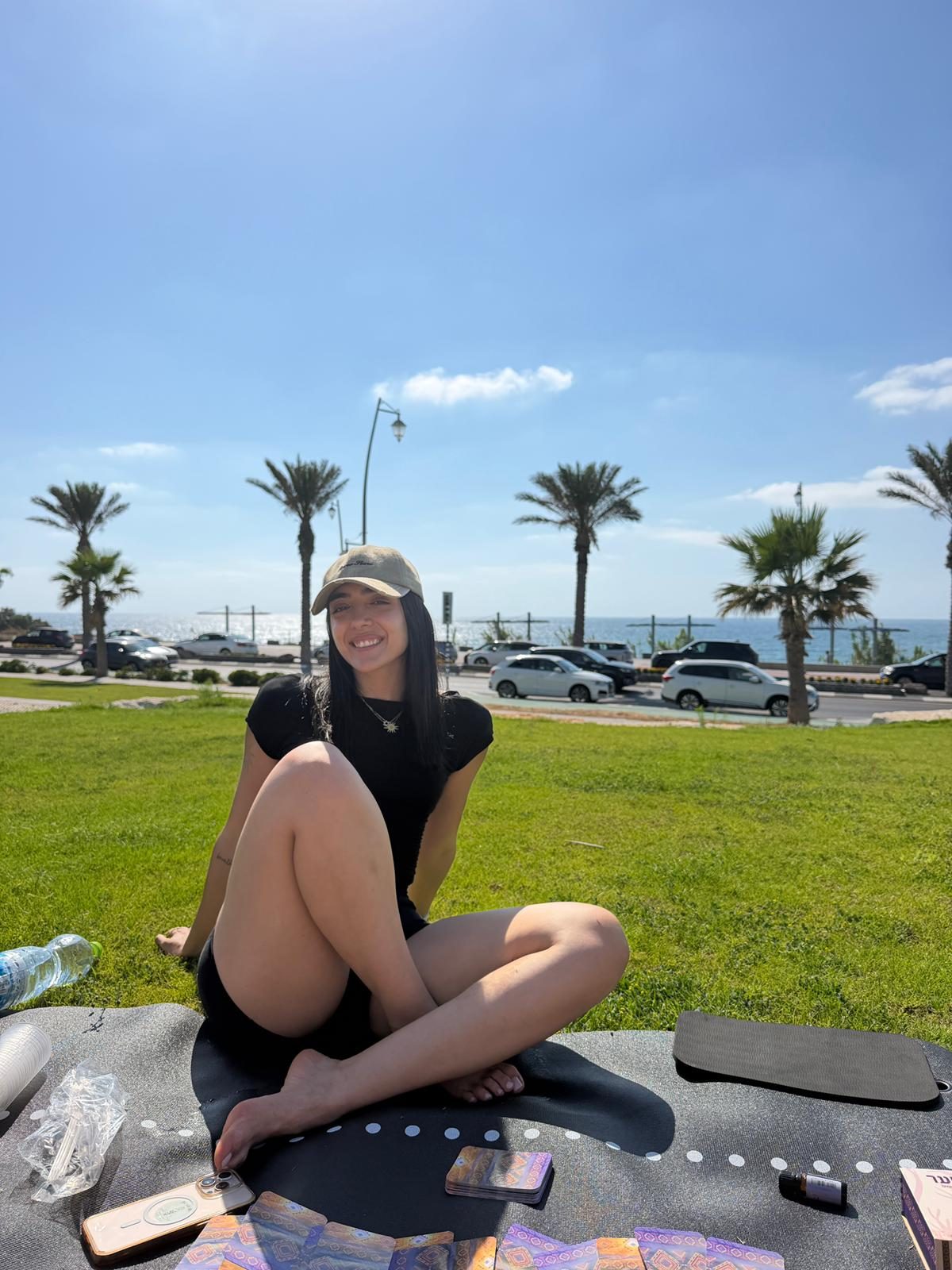 שירה דיין יושבת על מזרן על הדשא מול הים, קלפים פרוסים לפניה